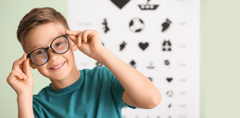 A young boy adjusting his glasses at the eye doctorโs office, stood in front of a Snellen chart