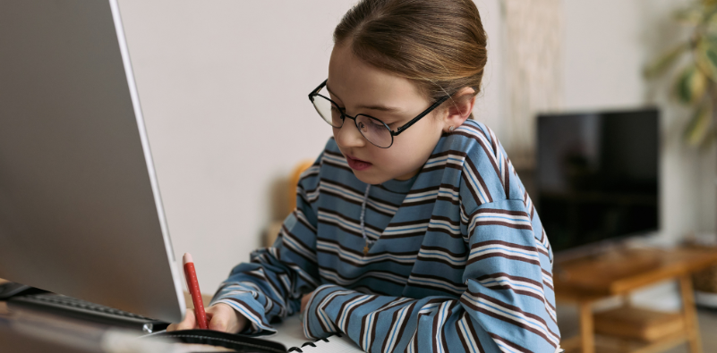 A young girl wearing glasses while writing beside a computer at home