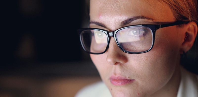 Close-up of a woman wearing black glasses while looking at a digital screen.