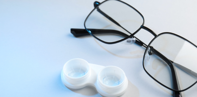 Black-framed glasses and an open contact lens case on a white surface.