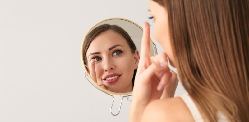 Woman applying a contact lens while looking into a handheld mirror.
